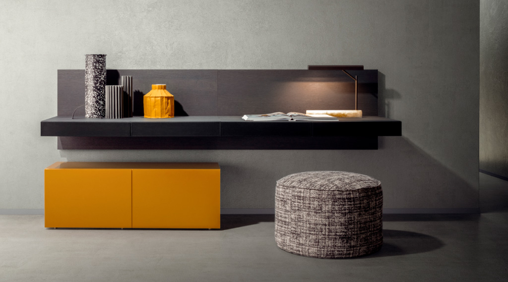 Consola moderna suspendida con acabado oscuro, decorada con jarrones y lámpara integrada, acompañada de un módulo bajo en color mostaza y un puf textil en primer plano.