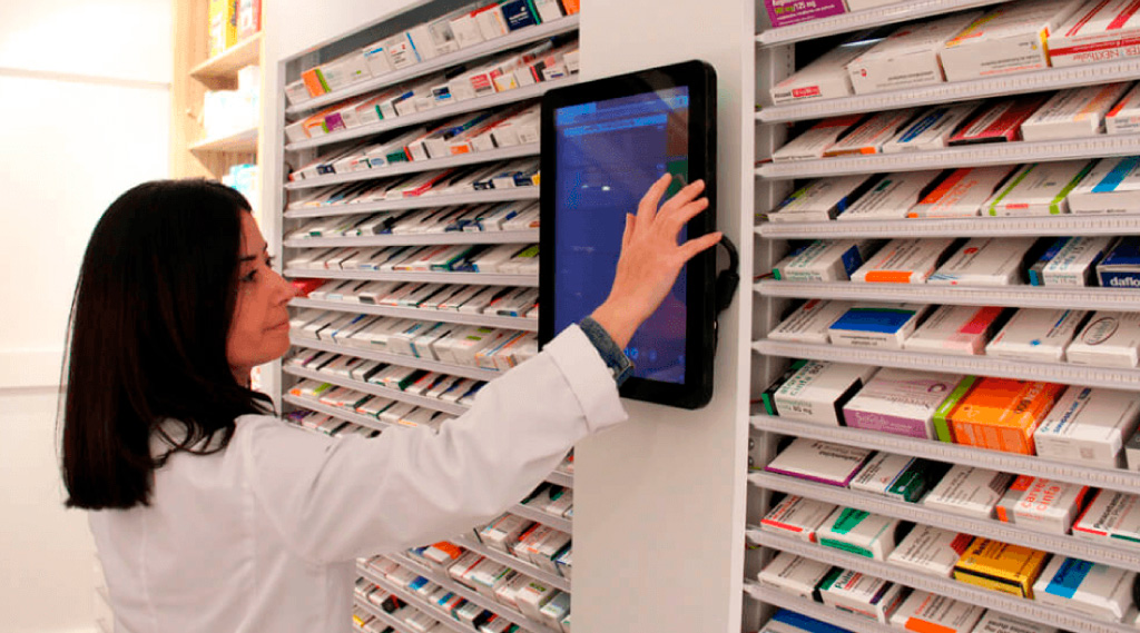 Farmacéutica utilizando una pantalla táctil para gestionar medicamentos frente a estanterías organizadas con cajas de productos farmacéuticos.