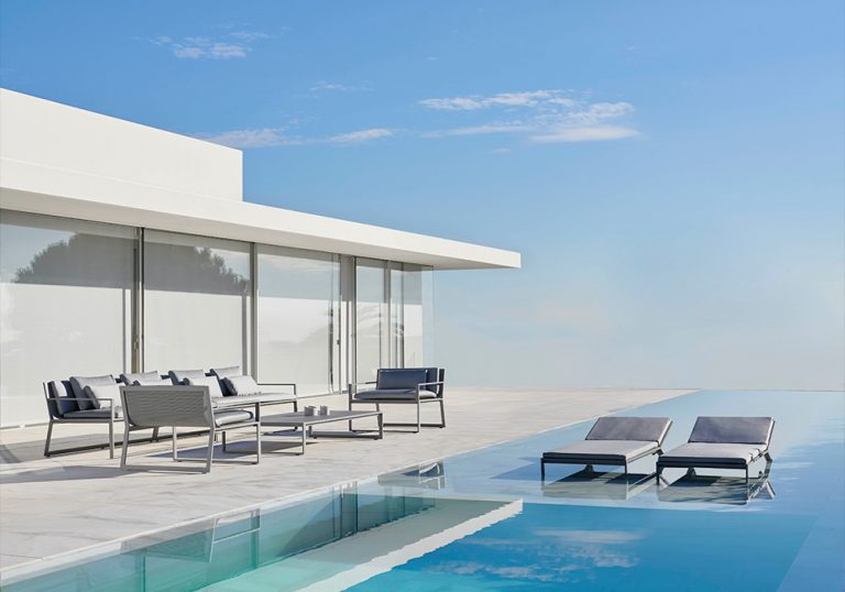 Terraza moderna con piscina infinita junto a vivienda de arquitectura minimalista, mobiliario exterior en tonos neutros y tumbonas parcialmente sumergidas frente a un paisaje abierto bajo cielo despejado