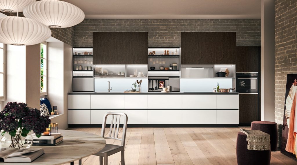 Cocina contemporánea con mobiliario en tonos blancos y madera oscura, estanterías abiertas iluminadas y pared de ladrillo visto en un espacio cálido y elegante.