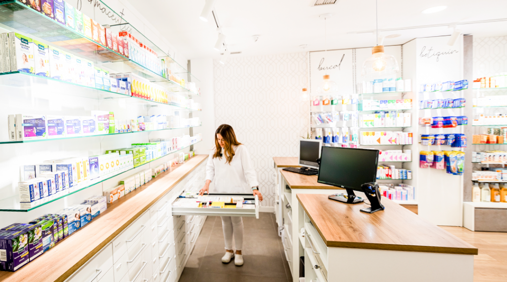 Farmacia moderna reformada con estanterías iluminadas de vidrio, mostrador de madera, zona de atención al cliente y profesional organizando productos en cajones, ejemplo de espacio optimizado para reformar farmacias con diseño funcional y luminoso.