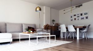 Salón moderno con sofá en tonos neutros, mesa de centro minimalista y zona de comedor integrada con mesa de madera, sillas blancas y lámparas decorativas.