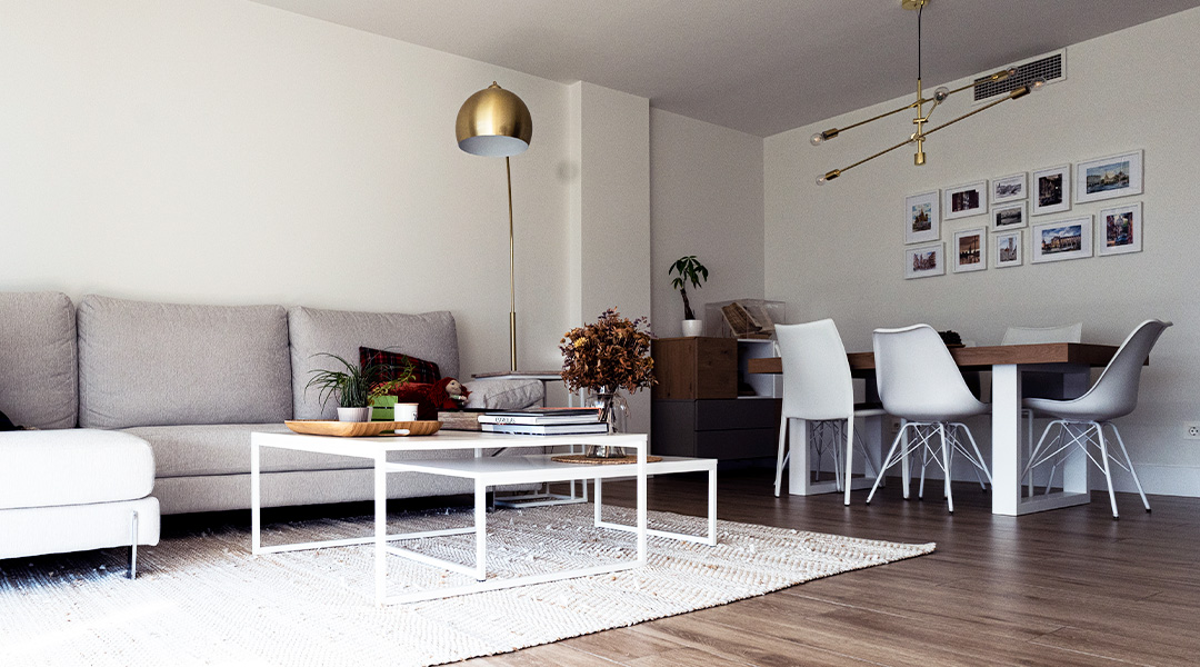 Salón moderno con sofá en tonos neutros, mesa de centro minimalista y zona de comedor integrada con mesa de madera, sillas blancas y lámparas decorativas.