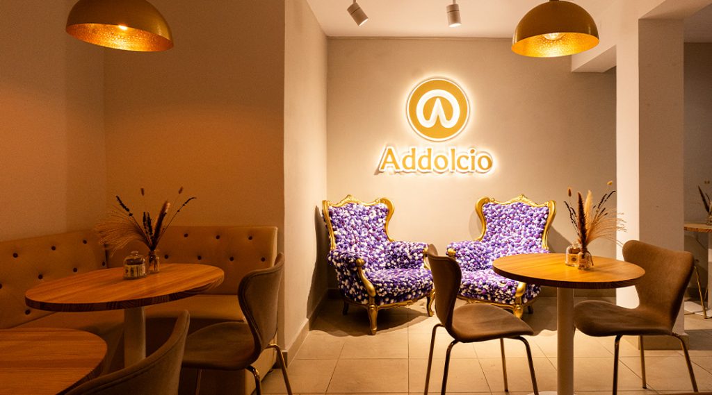 Interior de cafetería acogedora con mesas de madera, iluminación cálida, sillones decorativos y logotipo iluminado en la pared.