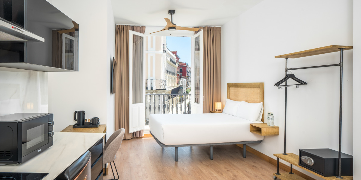 Habitación de hotel con cama, escritorio y balcón abierto con vistas urbanas, diseño moderno y funcional con luz natural