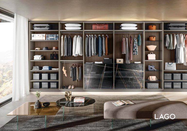 Vestidor amplio con armario empotrado abierto, estanterías organizadas con ropa colgada, cajas y accesorios, isla central con cajones y banco tapizado frente a mesa de centro moderna