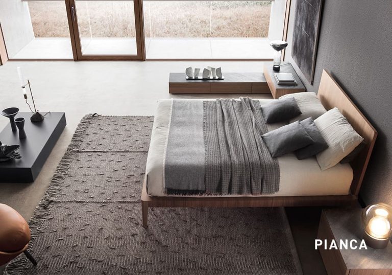 Dormitorio contemporáneo con cama de madera y textiles en tonos grises, alfombra amplia, mesillas bajas con objetos decorativos y ventanal con vistas al exterior que aporta luz