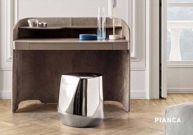 Tocador moderno de madera con cajón integrado y superficie superior con objetos decorativos, acompañado de taburete metálico brillante sobre suelo de parquet claro