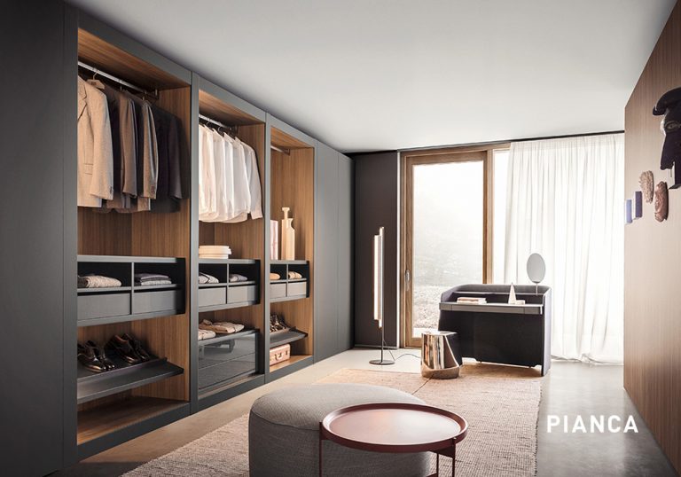 Vestidor moderno con armarios abiertos de madera y gris, ropa colgada y doblada, zona de tocador junto a ventana con cortinas claras y banco tapizado en el centro