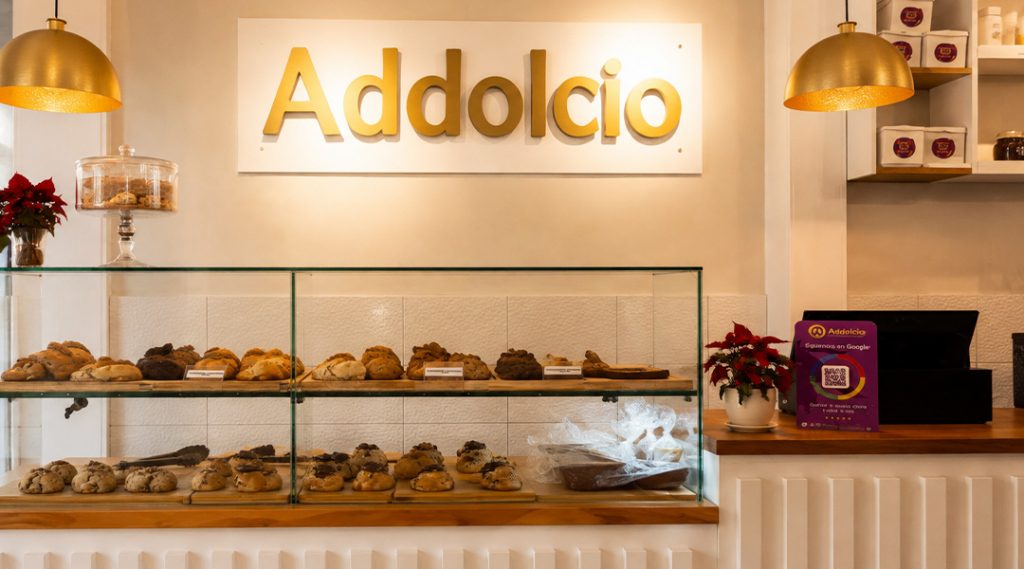 Bakery display case with two glass shelves of cookies and pastries, warm lighting, and an 'Addolcio' sign above.